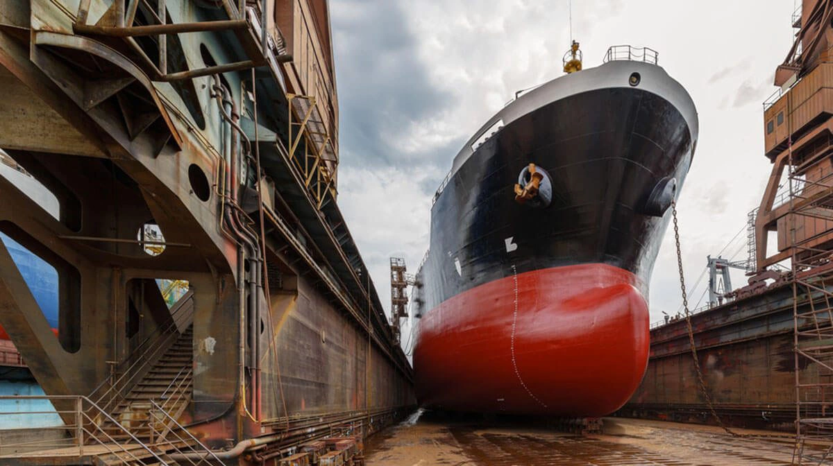 Large ship in a dry dock, indicating it is undergoing maintenance or repair. The ship's hull, painted in black and red with a visible waterline, towers over the dock, showing the scale and complexity of maritime vessels.