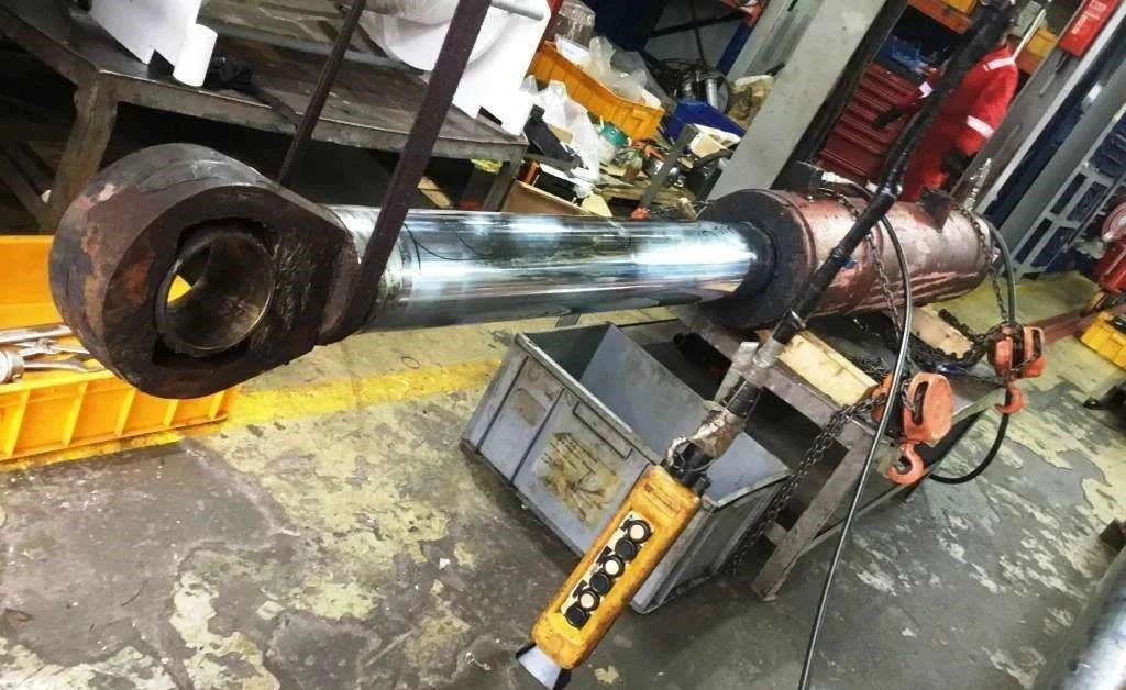 A large industrial hydraulic cylinder undergoing maintenance or repair on a workbench, with visible steel rods, heavy flanges, and hydraulic lines, highlighting the robust equipment used in heavy machinery